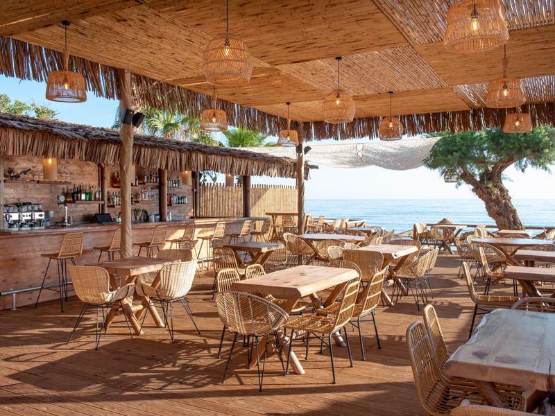 Open beach restaurant with wooden furniture, sea view, and tree in the background.