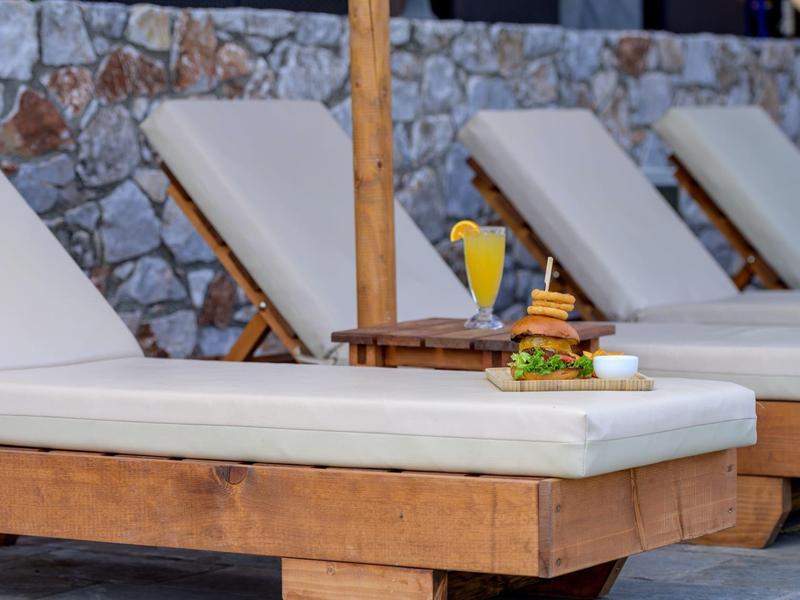 Lounge chair with a burger and drink beside a pool with a stone wall background.