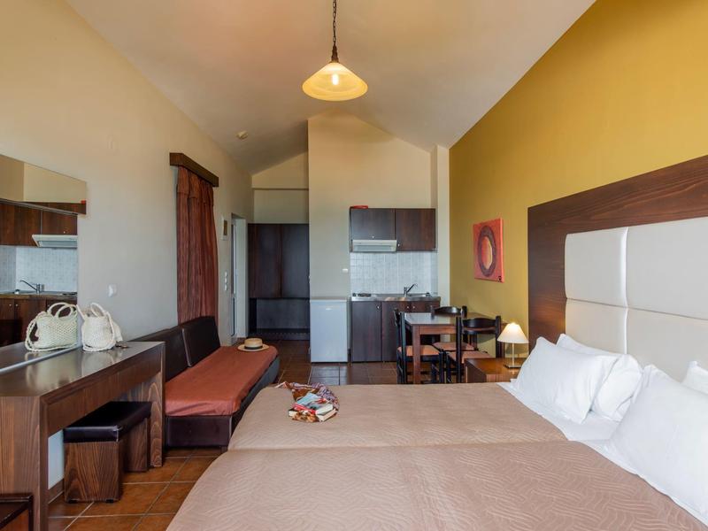 Hotel room with double bed, desk, bench, and small dining area in muted tones.