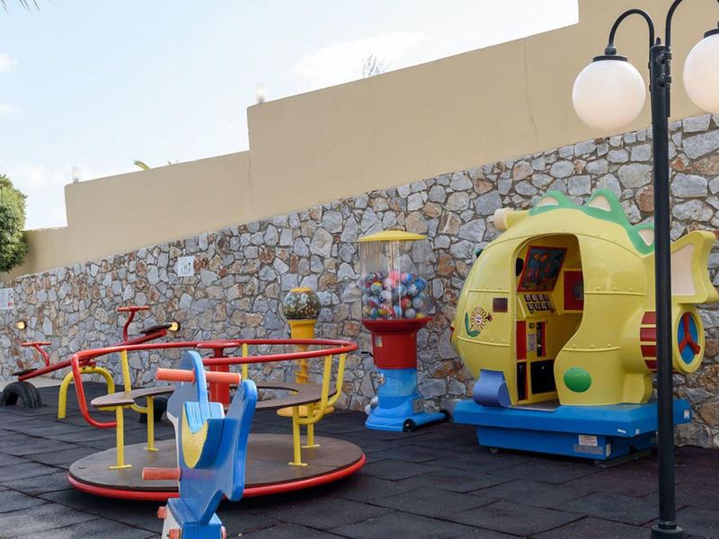 Colorful carousel and play equipment in a paved yard in front of a stone wall.