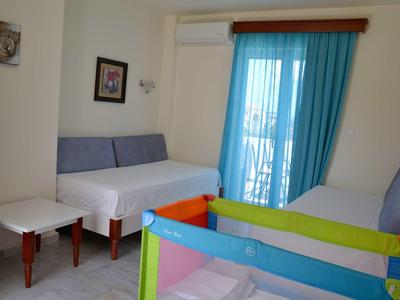 Kleine hotelkamer met twee bedden, een kinderbedje en blauwe gordijnen bij de balkondeur.