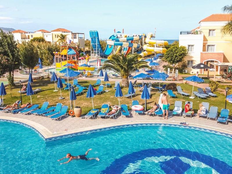 Groot hotelzwembad met blauwe ligbedden en parasols bij resortgebouwen op een zonnige dag.