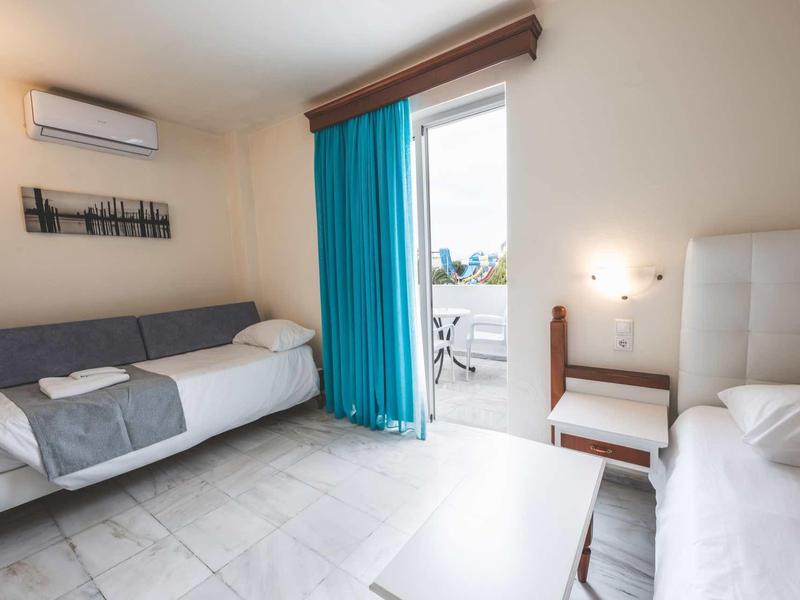 Lichte, schone hotelkamer met twee bedden, blauwe gordijnen en toegang tot balkon met zeezicht.