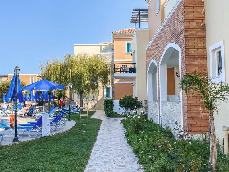 Pad langs een hotel met zwembad, ligstoelen en blauwe parasols onder een heldere hemel.
