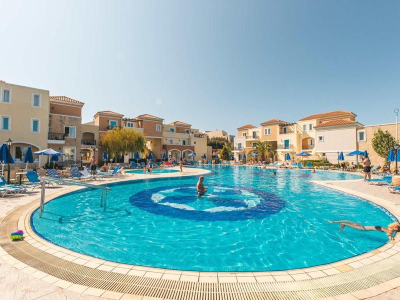 Groot rond zwembad met ligstoelen en mediterrane gebouwen op de achtergrond bij heldere lucht.