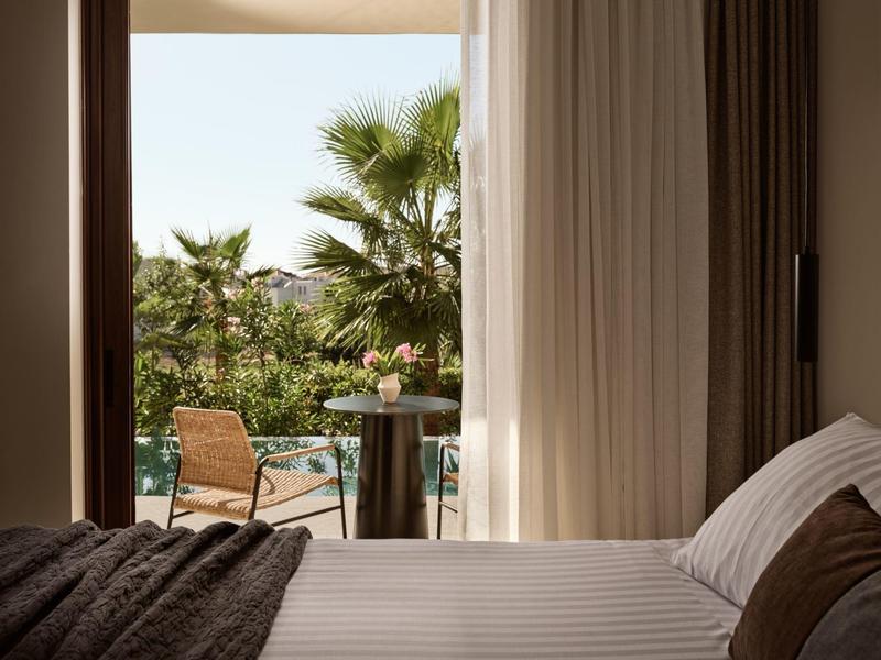 Vue depuis une chambre avec rideau ouvert sur une terrasse avec des palmiers et des chaises.