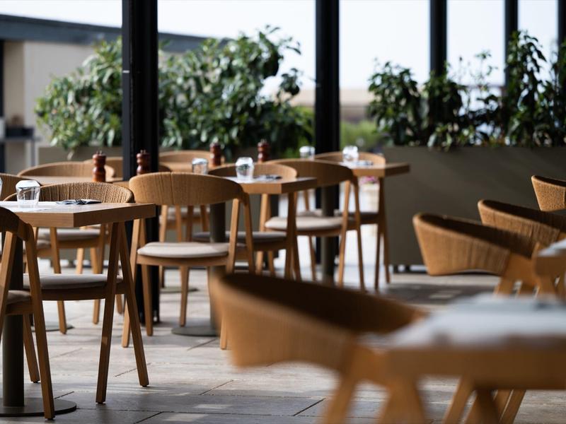 Salle à manger vide avec tables en bois, chaises et grandes fenêtres avec plantes dehors.