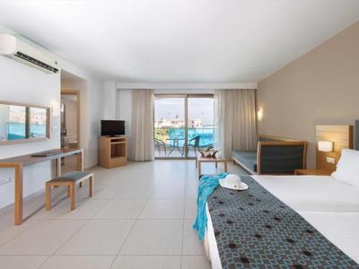 Bright hotel room with bed, TV, and balcony overlooking the pool area.