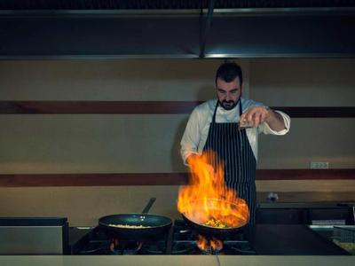 Chef wearing apron tossing flaming pan in professional kitchen.