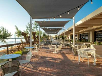 Überdachte Terrasse mit Tischen und Stühlen, Blick aufs Wasser und Palmen an einem Hotel.