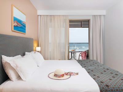 Modernes Hotelzimmer mit Doppelbett, Blick auf Meer, hellem Vorhang und dekorativem Bild an der Wand.