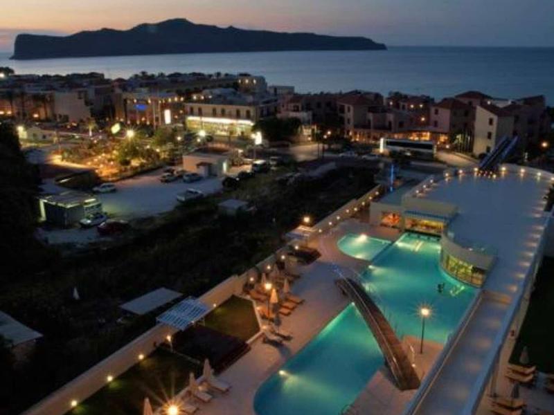 Evening view of a lit hotel pool area with sea and island in the background.