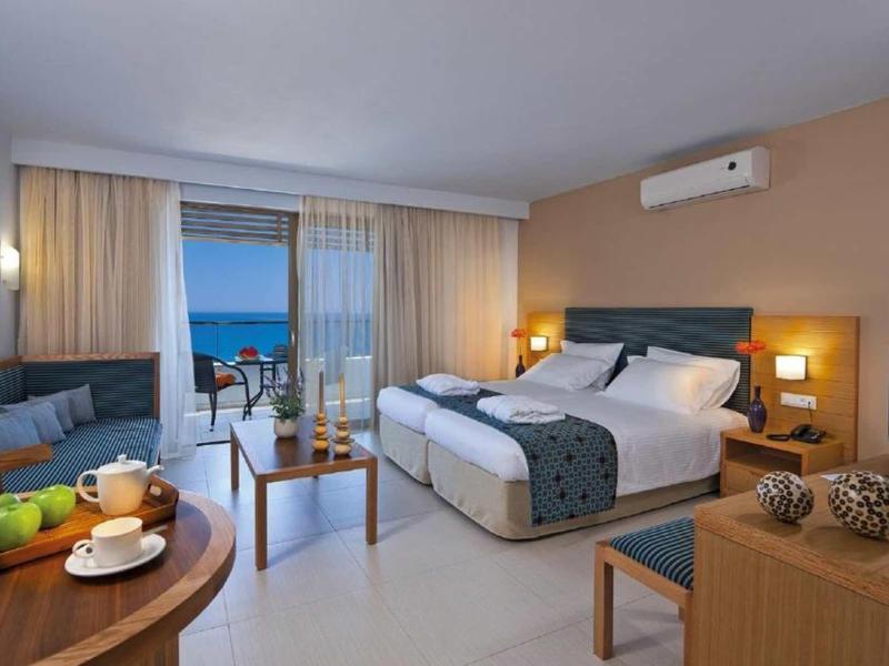 Modern hotel room with double bed, sofa, table, and balcony overlooking the sea.