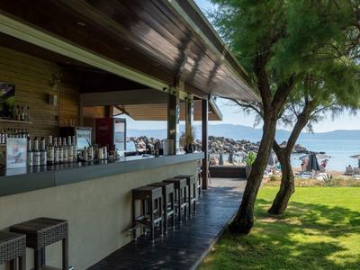 Offene Strandbar mit Barhockern, seitlich Bäume und Strand mit Sonnenschirmen und Menschen.