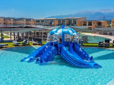 Venkovní bazén s modrými skluzavkami a lehátky pod jasnou oblohou v letovisku.