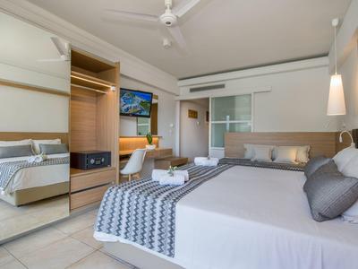 Prosvětlený hotelový pokoj s velkou postelí, stropním ventilátorem a televizí na zdi.