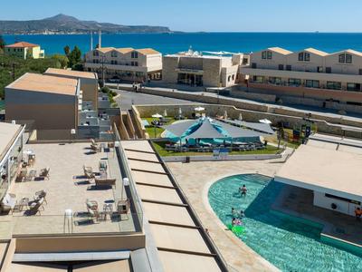 Blick auf ein Hotel mit Pool, Terrassenbereichen und Meer im Hintergrund an einem sonnigen Tag.