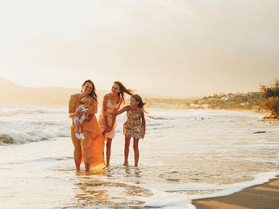 Tři ženy kráčejí po pláži v mělké vodě při západu slunce.