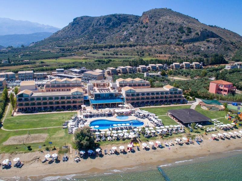 Velký hotel na pláži s několika bazény, lehátky a horským pozadím pod jasnou oblohou.