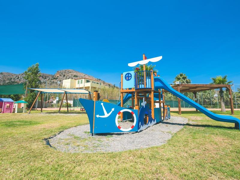 Barevné hřiště s lezecími konstrukcemi a skluzavkou na zelené trávě pod jasnou modrou oblohou.