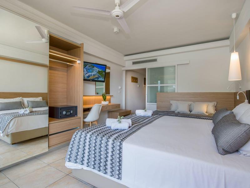 Prosvětlený hotelový pokoj s velkou postelí, stropním ventilátorem a televizí na zdi.