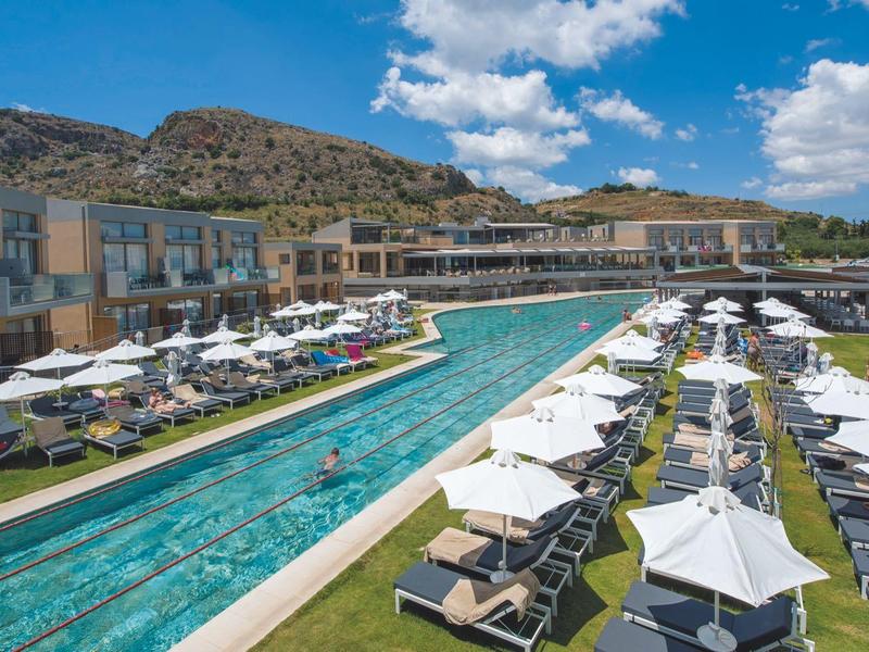 Moderní hotel se dvěma dlouhými bazény, lehátky a slunečníky pod modrou oblohou.