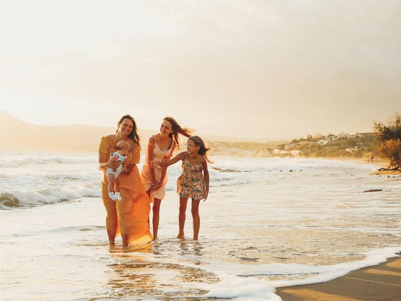 Tři ženy kráčejí po pláži v mělké vodě při západu slunce.