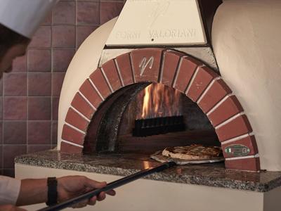 Ein Koch holt eine Pizza aus einem gemauerten Holzofen mit brennendem Feuer.