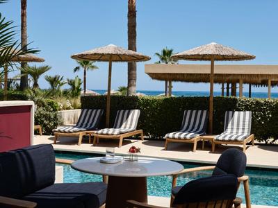 Poolbereich mit Sonnenliegen, Schirmen und mehrstöckigem Gebäude im Hintergrund bei klarem Himmel.