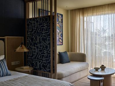 Modernes Hotelzimmer mit Bett, Sofa, Beistelltischen und großen Vorhängen am Fenster.