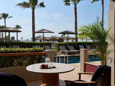 Blick auf einen Poolbereich mit Liegestühlen, Schirmen und Palmen an einem sonnigen Tag.