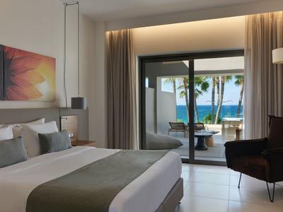 Habitación de hotel moderna con cama king, área de estar y vista al mar y piscina a través de grandes puertas de vidrio.