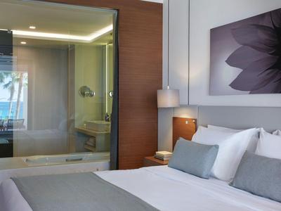 Habitación de hotel moderna con cama grande, almohadas grises y pared de vidrio al baño.