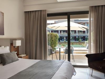 Habitación de hotel luminosa con cama doble, silla cómoda y vista a la terraza con piscina.