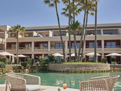 Ein Hotel mit einem runden Pool und zwei weißen Stühlen unter Palmen an einem sonnigen Tag.