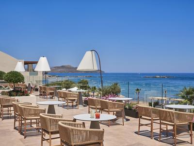 Terrasse mit Tischen und Sesseln bietet Blick auf das Meer und Sanddünen unter blauem Himmel.