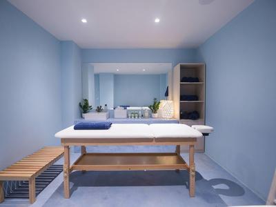 Kleiner Raum mit blauen Wänden, Handtuch auf Massagetisch, Holzbank und Spiegel an der Wand.