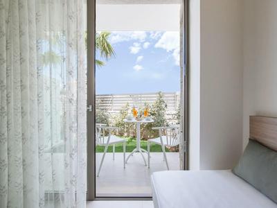 Blick durch ein Glasfenster auf Balkon mit weißen Möbeln, grüner Rasen und blauer Himmel.