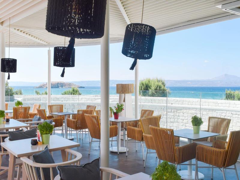 Modernes Strandrestaurant mit weißen Tischen, braunen Korbstühlen und Meerblick bei klarem Himmel.