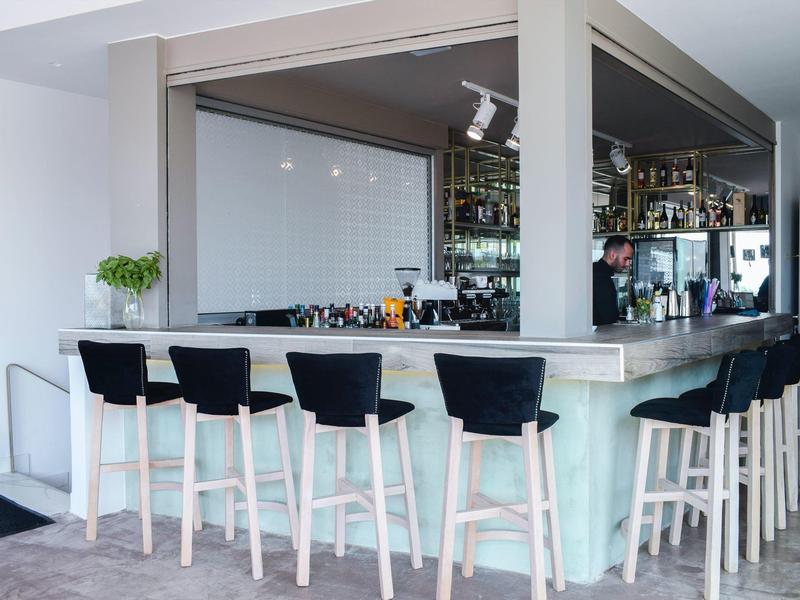 Modern eingerichtete Bar mit sechs schwarzen Hockern und Glasregalen im hellen Raum.