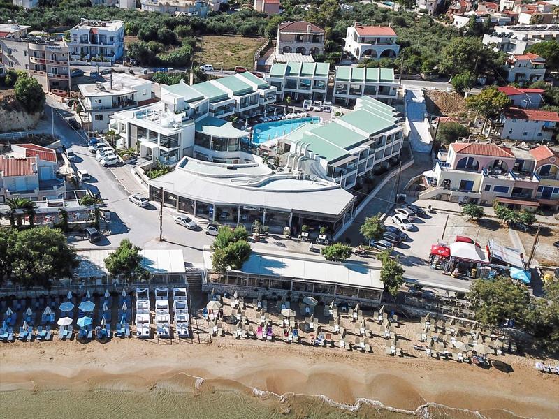 Luftaufnahme eines Hotelkomplexes mit Pool, Strand und Liegestühlen am Meer vor bewaldetem Hügel.