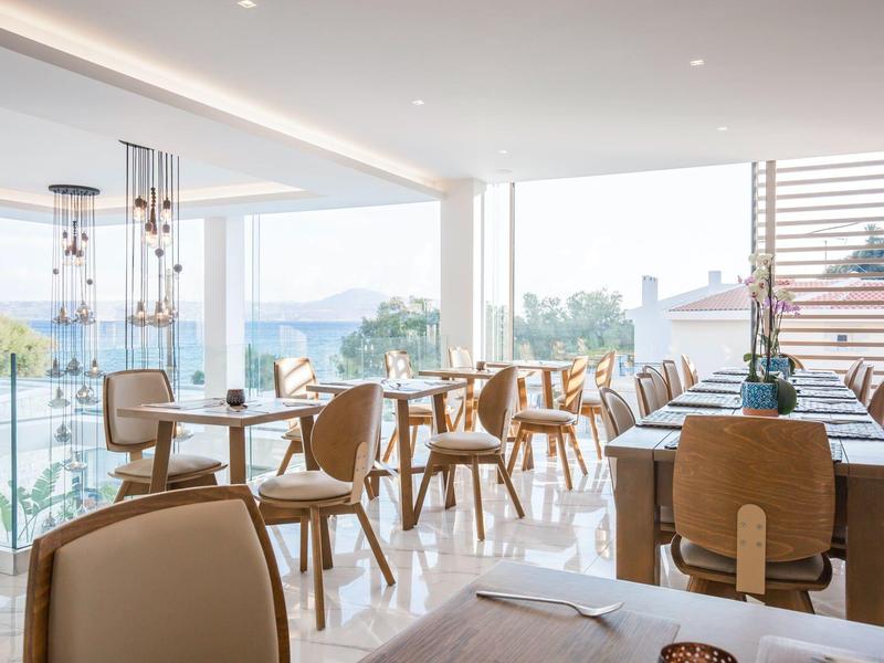 Helles, modernes Restaurant mit großen Fenstern, Holzmöbeln und Blick auf Wasser und Sonne.