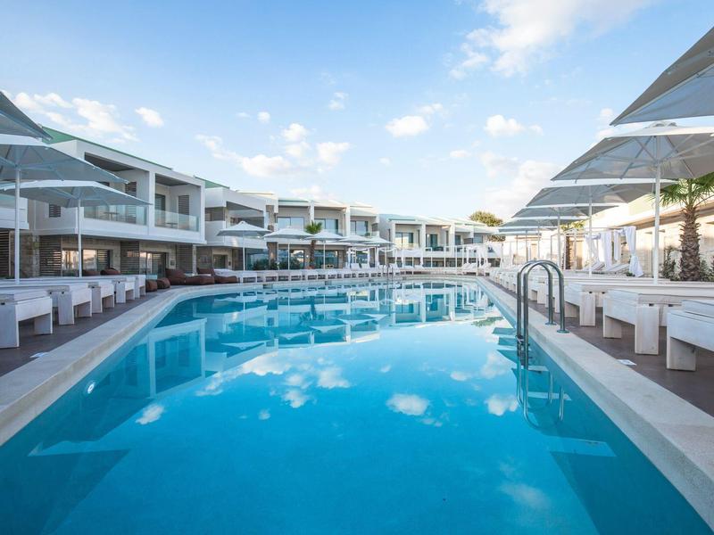 Großer blauer Pool mit weißen Liegen und modernen Gebäuden unter blauem Himmel.