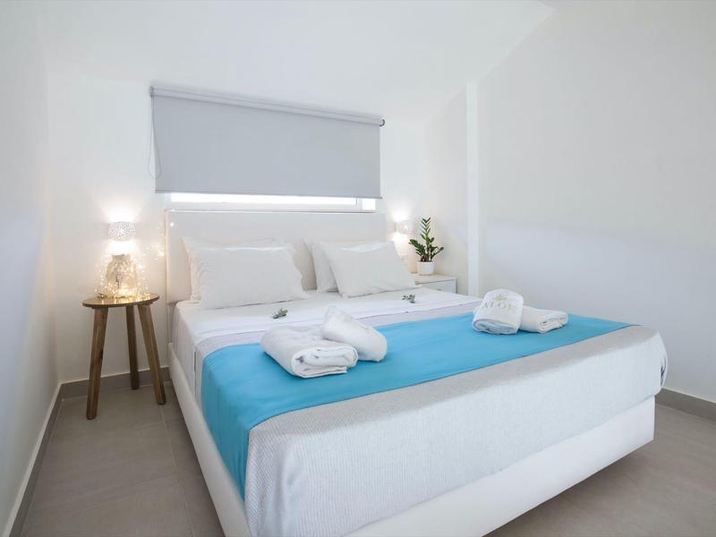 Weißes Schlafzimmer mit Doppelbett, blauer Tagesdecke und Handtüchern, seitlicher Lampe und Blume.