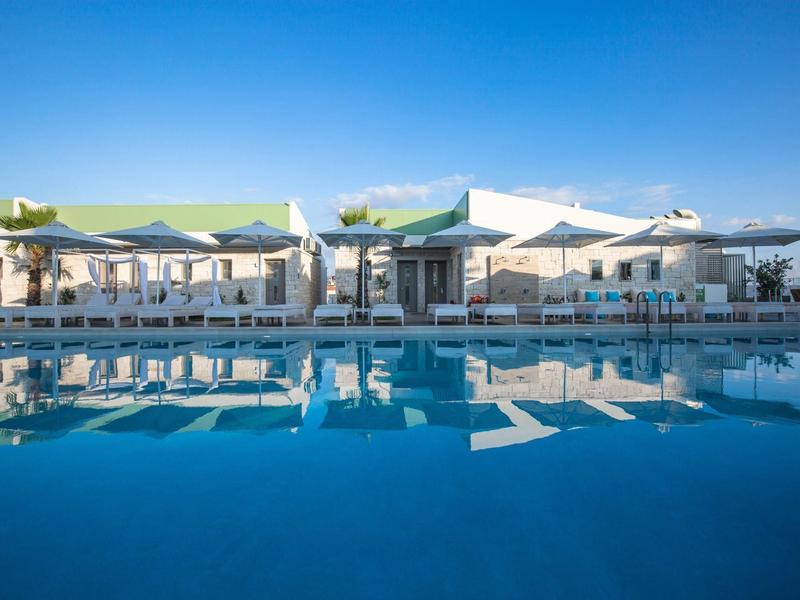 Großer Pool mit klar blauem Wasser vor weißem Gebäude mit grünen Dächern und Sonnenschirmen.