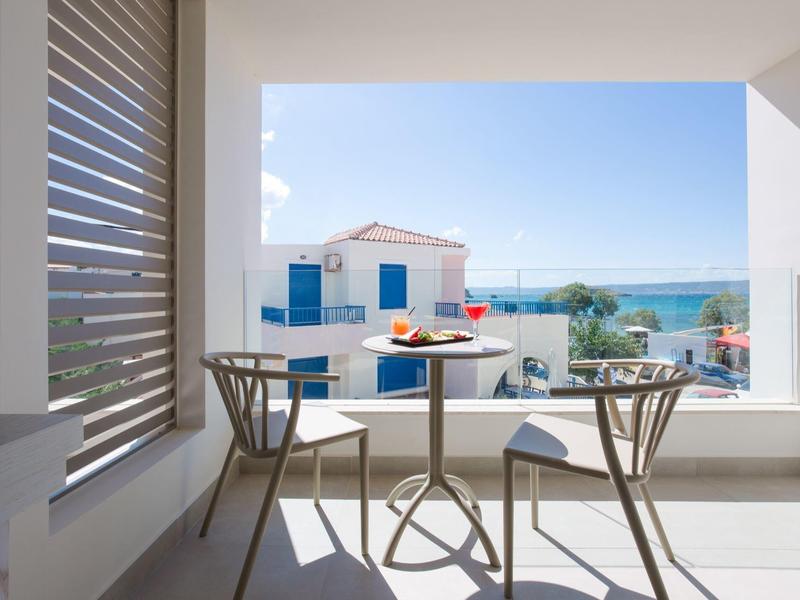 Heller Balkon mit Tisch, zwei Stühlen, Blick auf weiße Häuser und blaues Meer bei klarem Himmel.