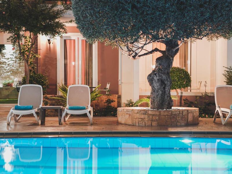 Verlicht zwembad met witte ligstoelen en een boom op een hotelterras.