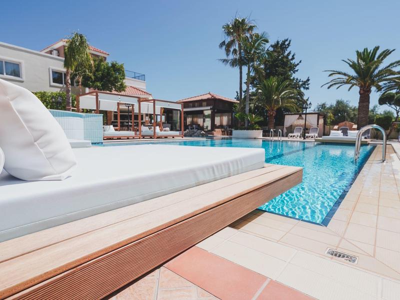 Modern hotelzwembad met witte ligbedden, palmbomen en parasols bij heldere lucht.