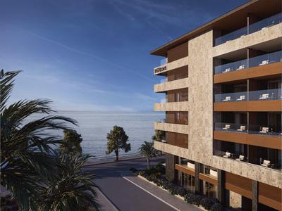 Moderní hotelová budova s balkony a palmami u moře pod modrou oblohou.