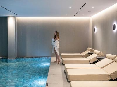 Femme debout près d'une piscine intérieure avec chaises longues et éclairage doux.